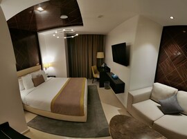 A modern hotel room with a curved bed, a flat-screen TV, a sofa, and a desk with a lamp.