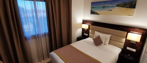 A hotel room with a large bed, two bedside tables with lamps, a painting of a beach scene, and a view through sheer curtains.