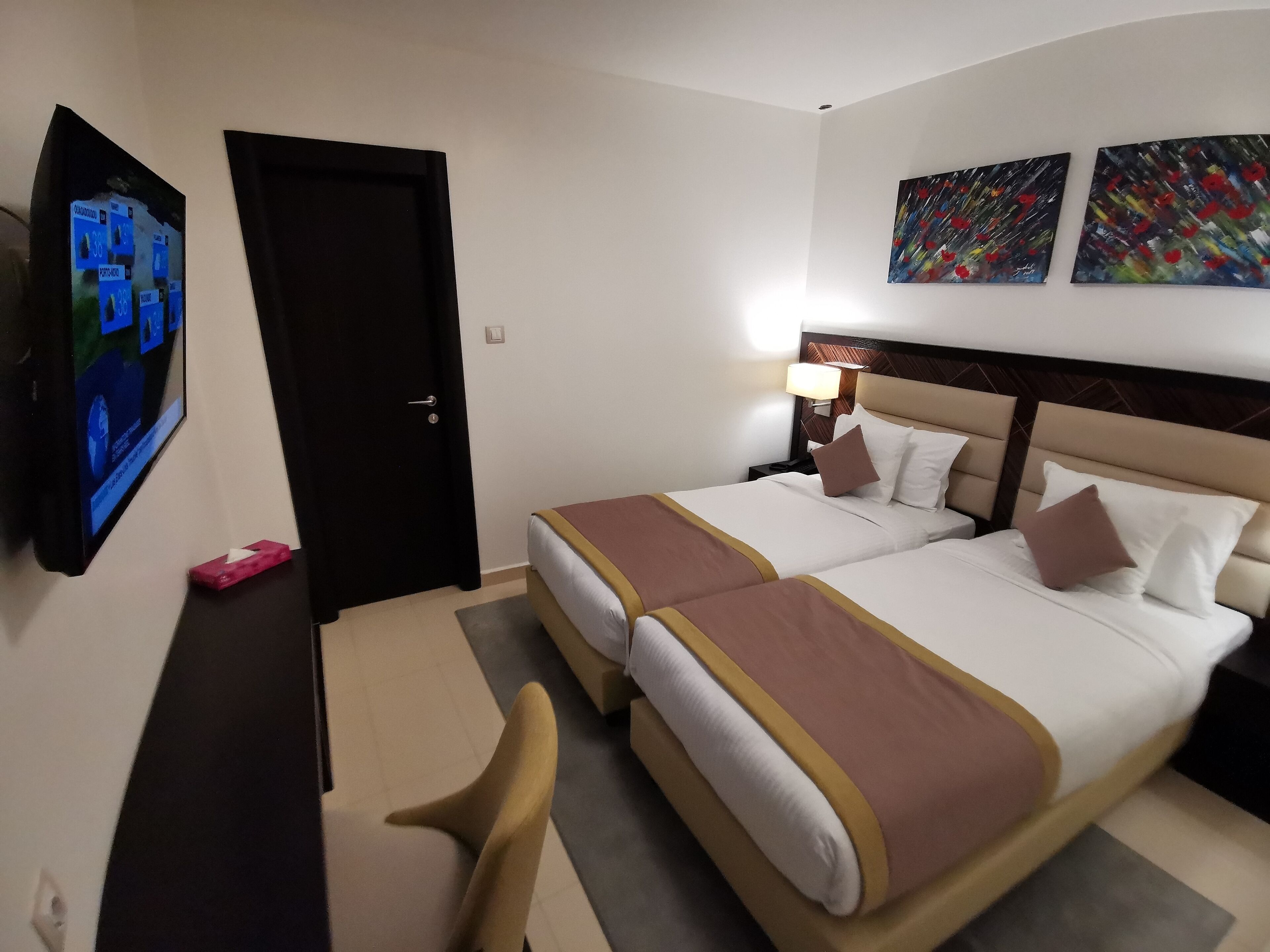 Een hotelkamer met twee bedden, een aan de muur bevestigde televisie, en twee schilderijen boven de bedden.