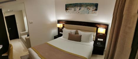 A hotel room with a large bed, two bedside lamps, a painting on the wall, and a door leading to another room.