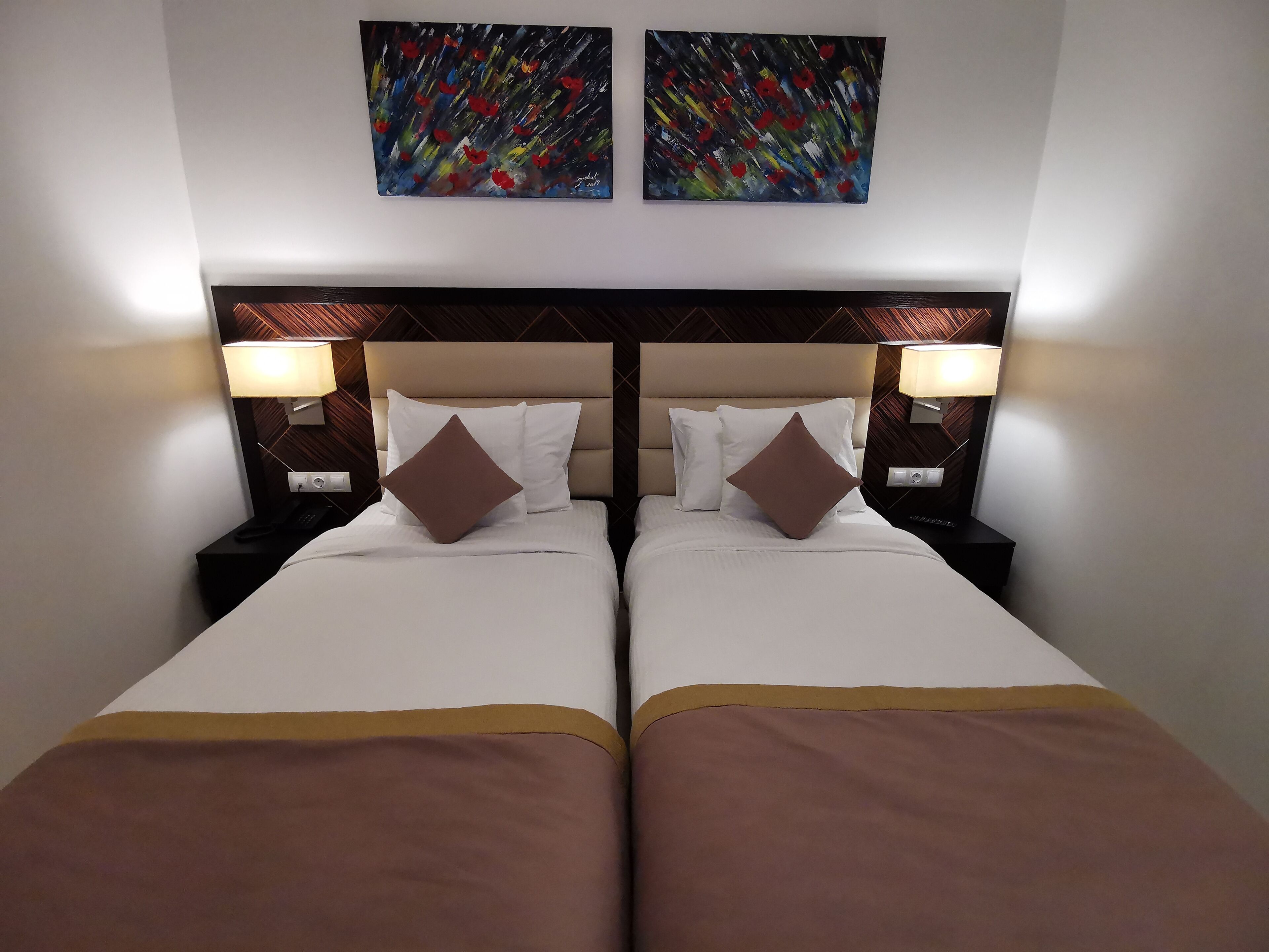 Habitación de hotel con dos camas, cada una con cubrecama marrón, almohadas blancas y cabeceros color beige. Hay dos lámparas de pared a cada lado de la cama.