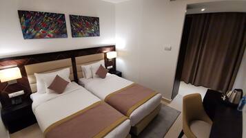 Quarto de hotel com duas camas, uma mesa, uma cadeira e duas pinturas abstratas na parede.