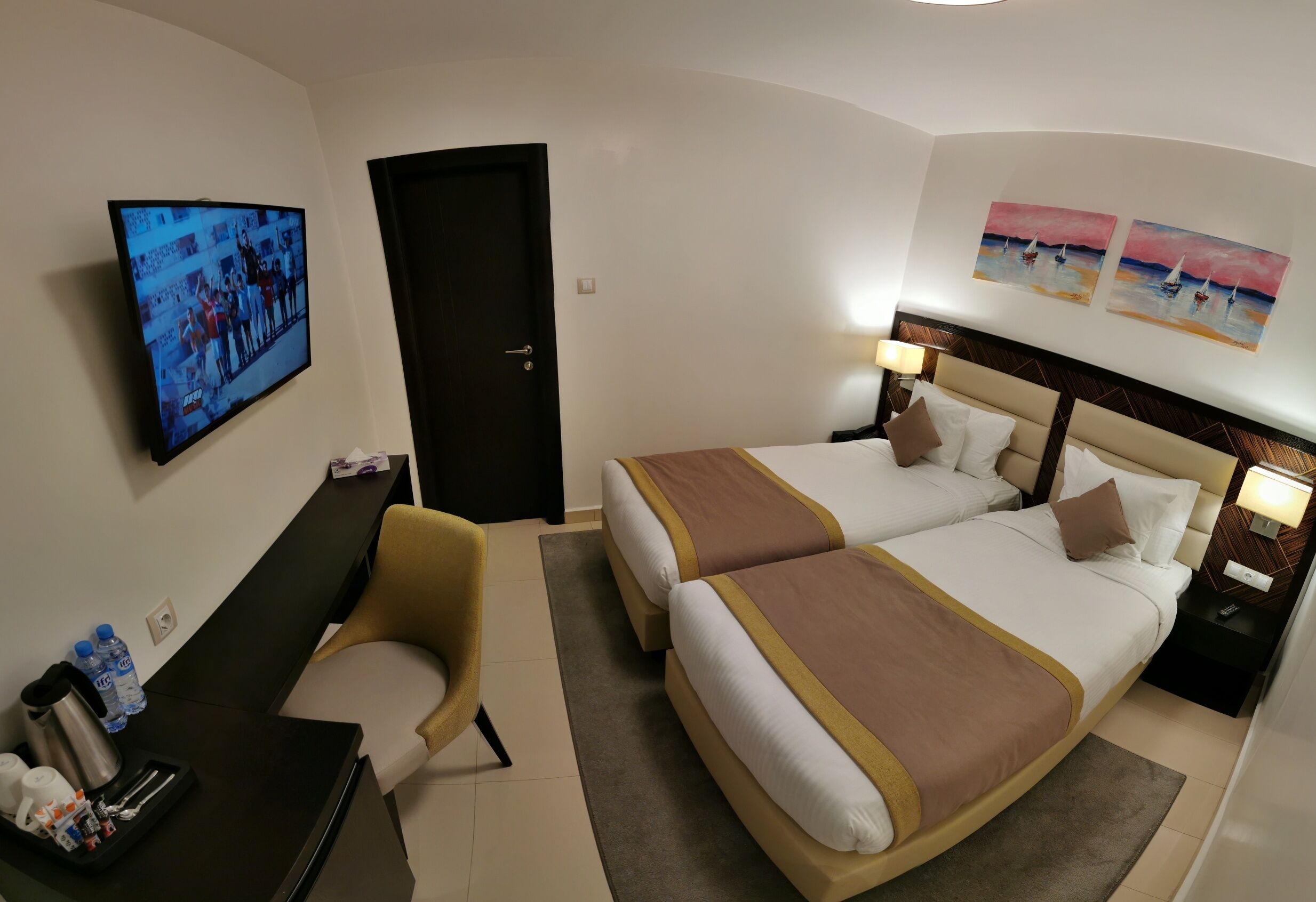 A hotel room with two beds, a desk, a chair, a TV, and artwork on the wall.