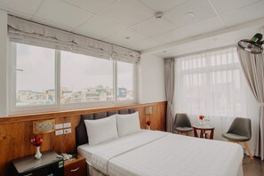 Une chambre d’hôtel avec un grand lit, deux fauteuils, une petite table et une vue sur la ville grâce à de grandes fenêtres.
