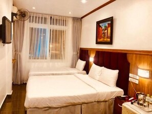Une chambre d’hôtel avec deux lits, une grande fenêtre avec des rideaux légers, un tableau au mur et une table de chevet avec une lampe et une bouilloire.