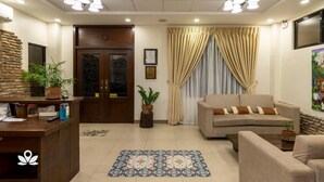 A well-lit living room with a sofa, a coffee table, and a reception desk. There are two potted plants, a wooden door, and a framed certificate on the wall.