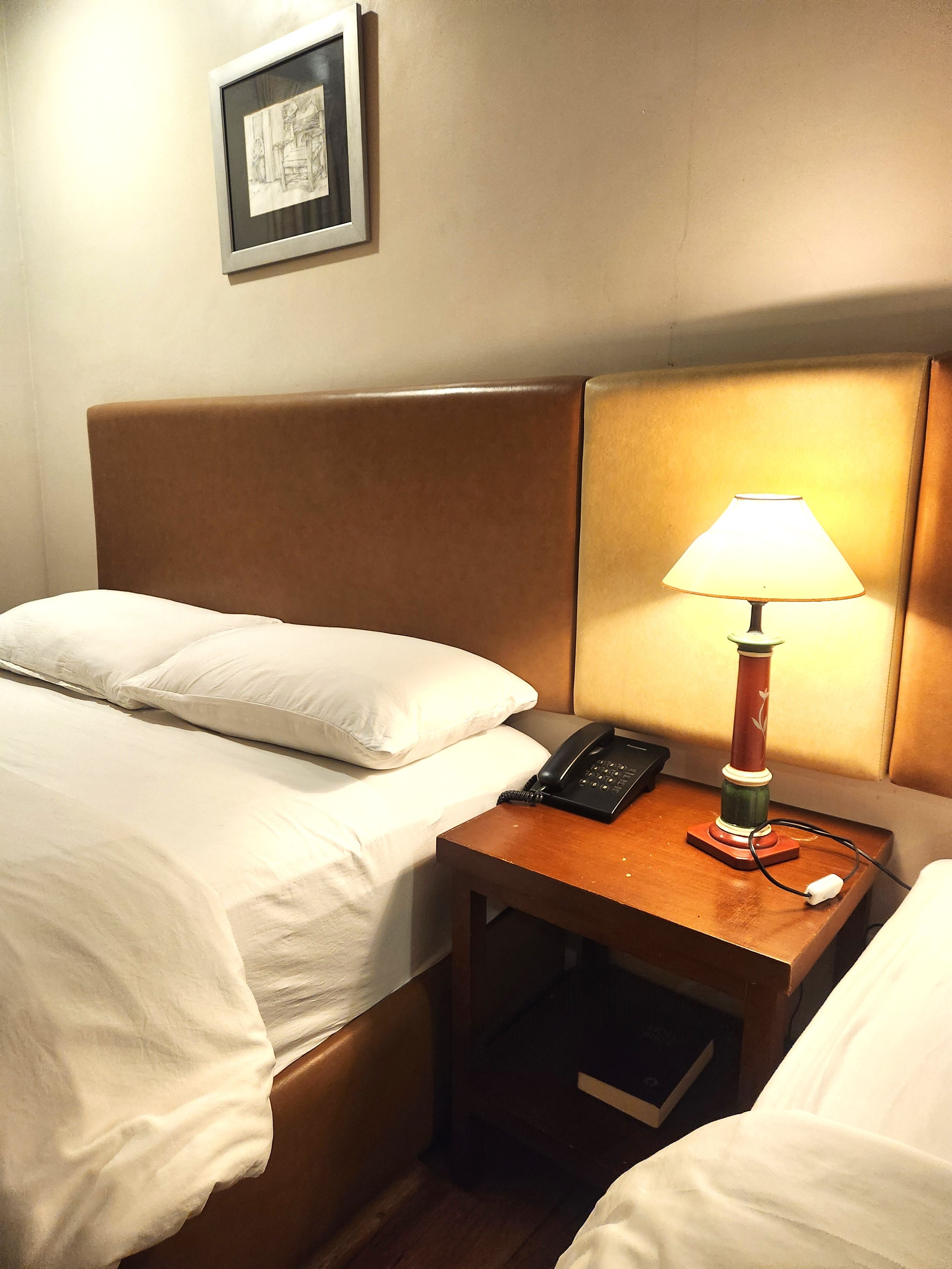 A hotel room with a bed, a bedside table with a lamp, a phone, and a framed picture on the wall.