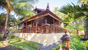 A traditional wooden house with a thatched roof, surrounded by a well-maintained garden with palm trees and a stone lantern. - Teluk Indah Beach & Pool Villa (Tejakula)