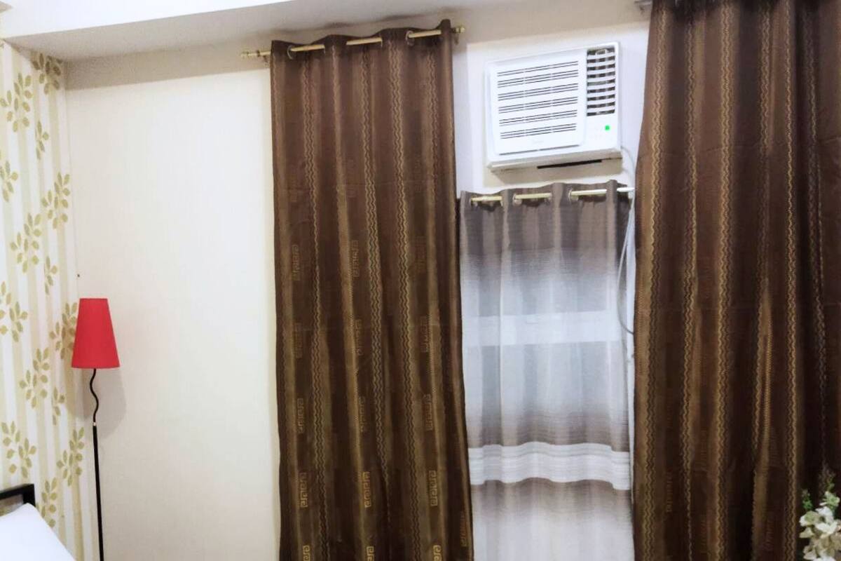 Una habitació d'hotel amb un llit, una làmpada vermella, cortines marrons, un aparell d'aire condicionat i una taula petita amb una planta.