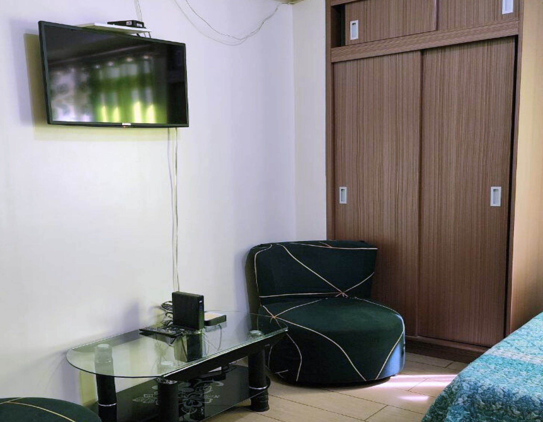Une petite pièce avec un téléviseur fixé au mur, une table en verre avec un ordinateur, un fauteuil recouvert d’un tissu vert et une armoire en bois.