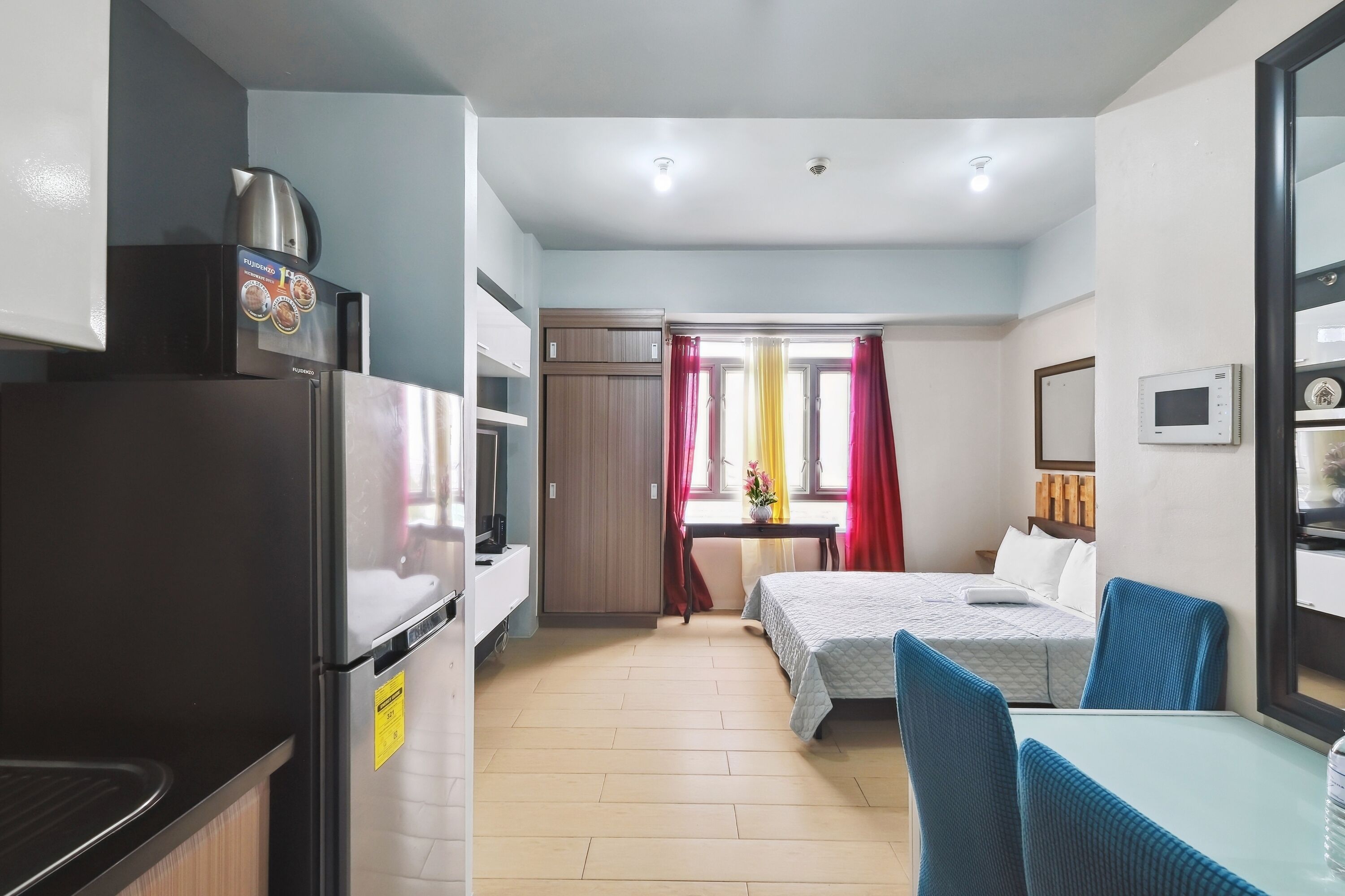 A compact hotel room with a kitchenette, a bed with a white duvet, a small table with two chairs, and a window with curtains.