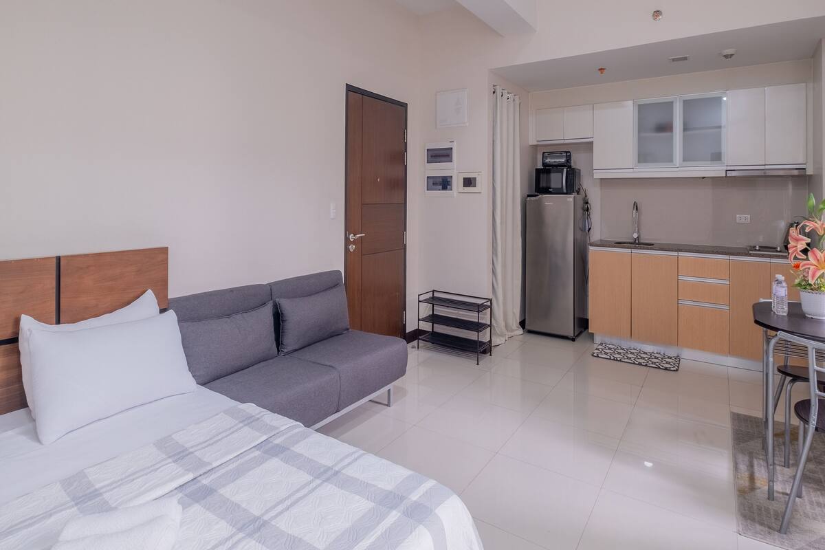 Habitació d'hotel moderna amb llit, sofà gris, petita cuina i zona de menjador.