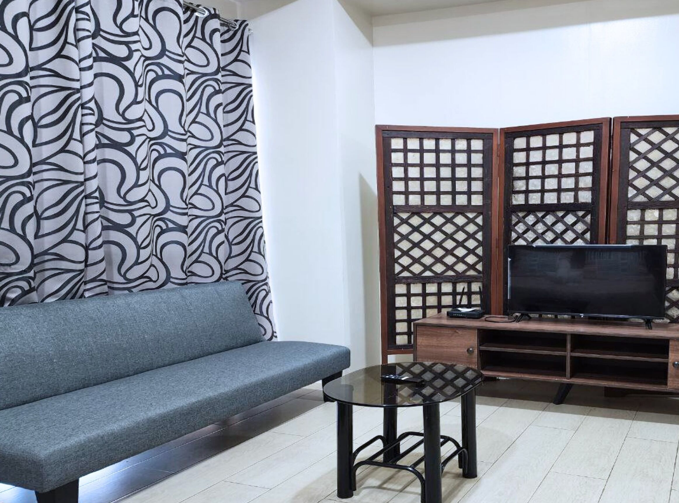 A living room with a grey sofa, a glass coffee table, a TV on a wooden stand, and a decorative screen with a geometric pattern.