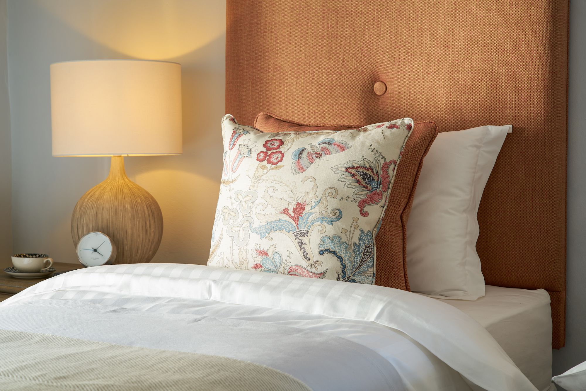A neatly made bed with a floral pillow, a plain pillow, and a textured headboard. A lamp with a round base and a white shade is on a bedside table.