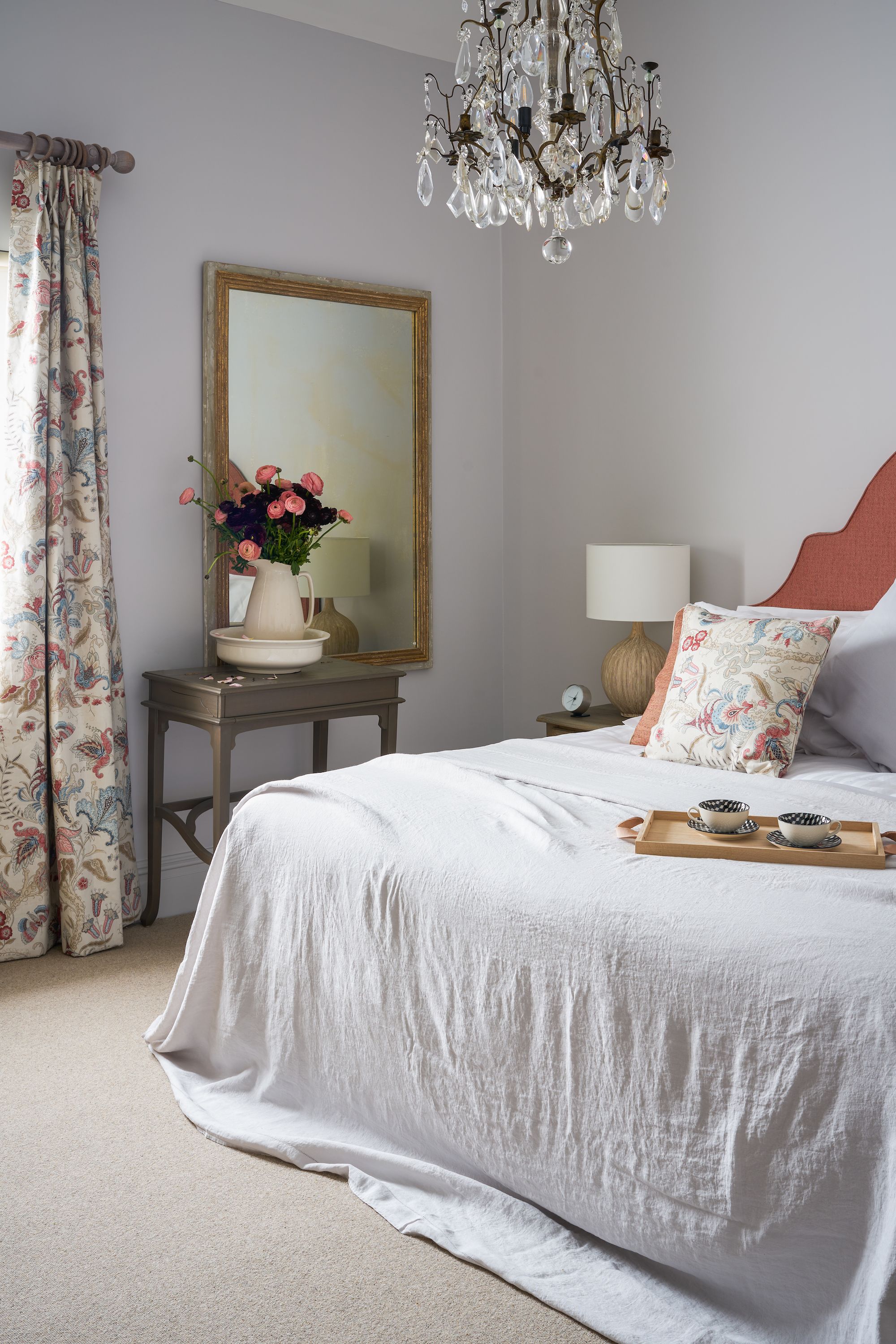 A bedroom with a large bed, a chandelier, a mirror, a bedside table with a lamp, and a tray with cups.