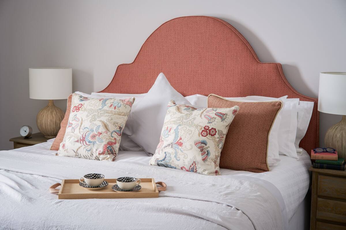 A neatly made bed with a pink headboard, two decorative pillows, a tray with two cups, and a bedside table with a lamp and books.