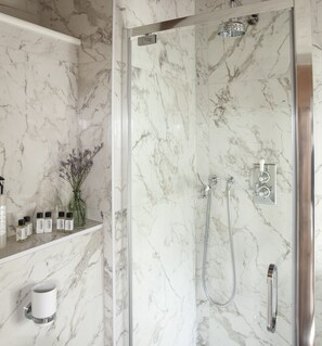 A bathroom with a marble shower enclosure, a glass door, and a shelf with toiletries.