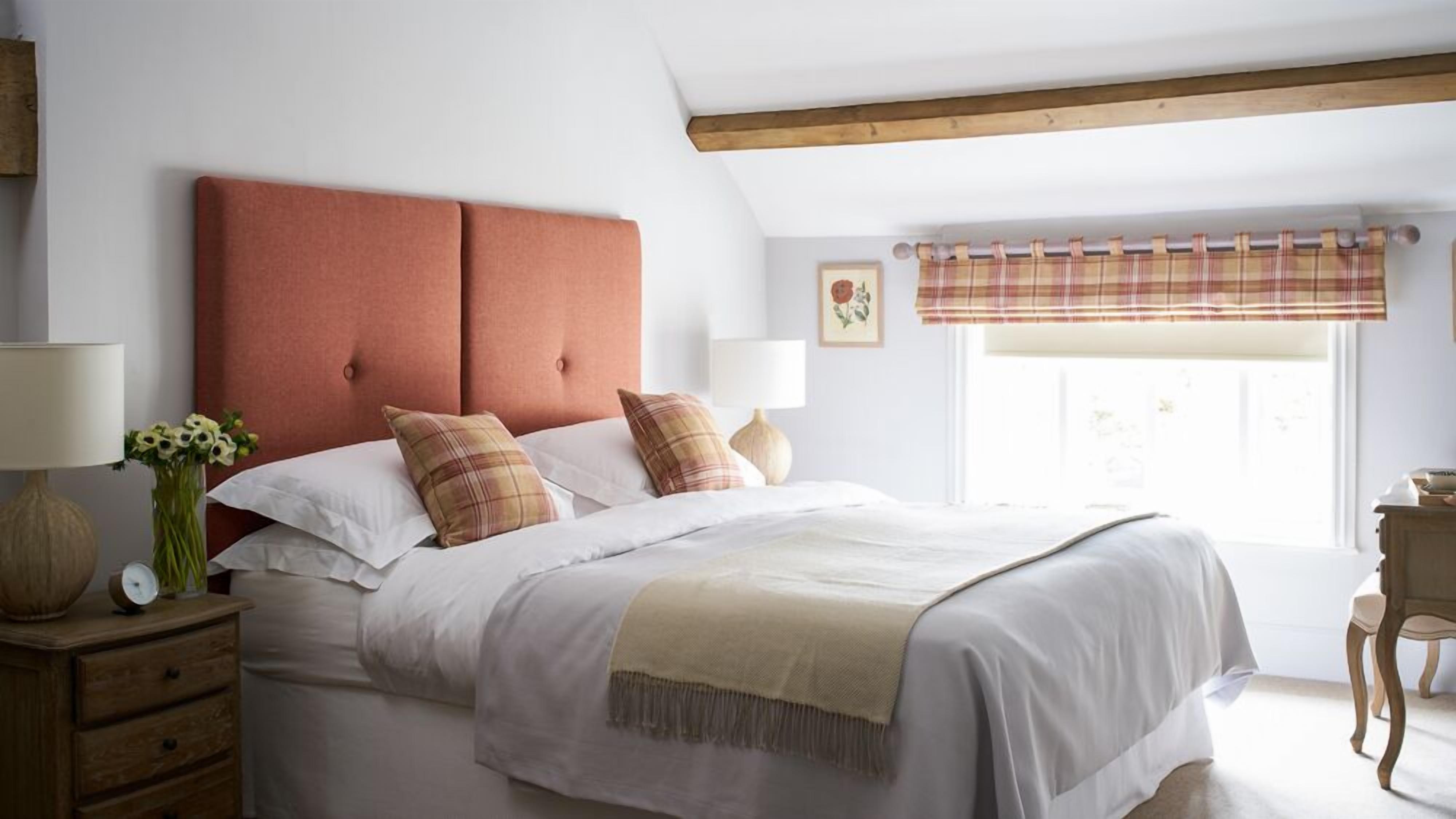 A neatly made bed with a red headboard, white bedding, and a beige throw blanket. There is a wooden nightstand with a lamp and a vase of flowers beside the bed. A window with a plaid curtain is visible in the background.