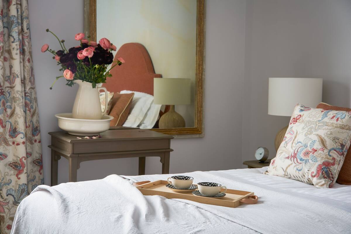 A neatly made bed with a tray of cups and saucers, a vase with flowers, a lamp, and a mirror on the wall.