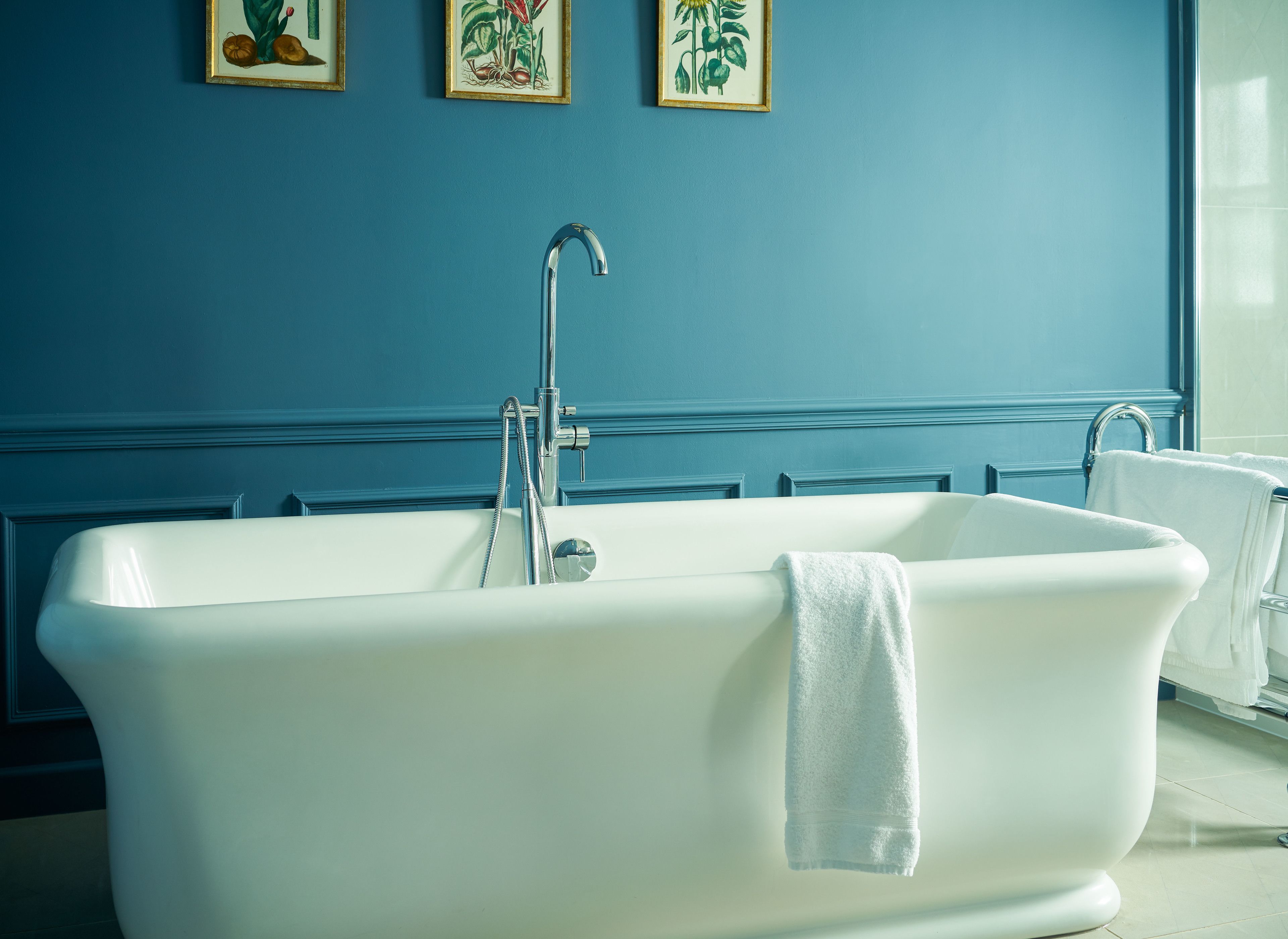 A white freestanding bathtub in a bathroom with blue walls and framed botanical prints.
