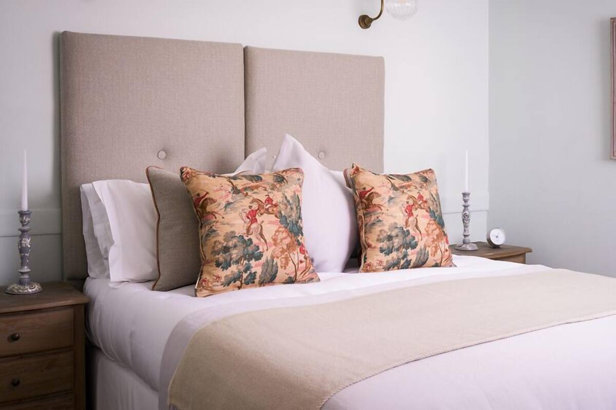 A neatly made bed with floral-patterned pillows, a beige headboard, and a bedside table with a clock and a candle.