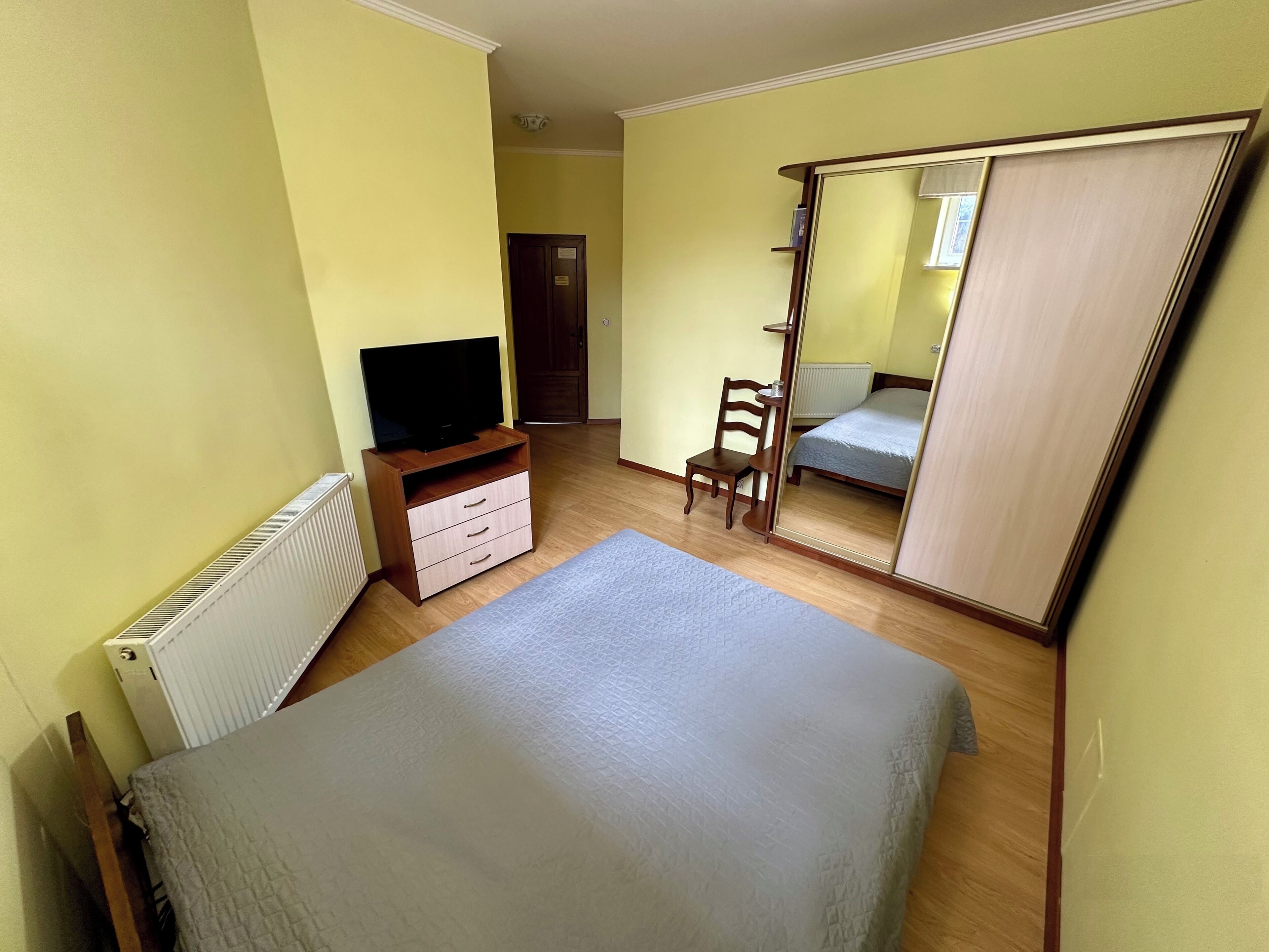Un dormitorio con una cama, un televisor sobre un mueble de madera, una silla, un espejo y un armario.