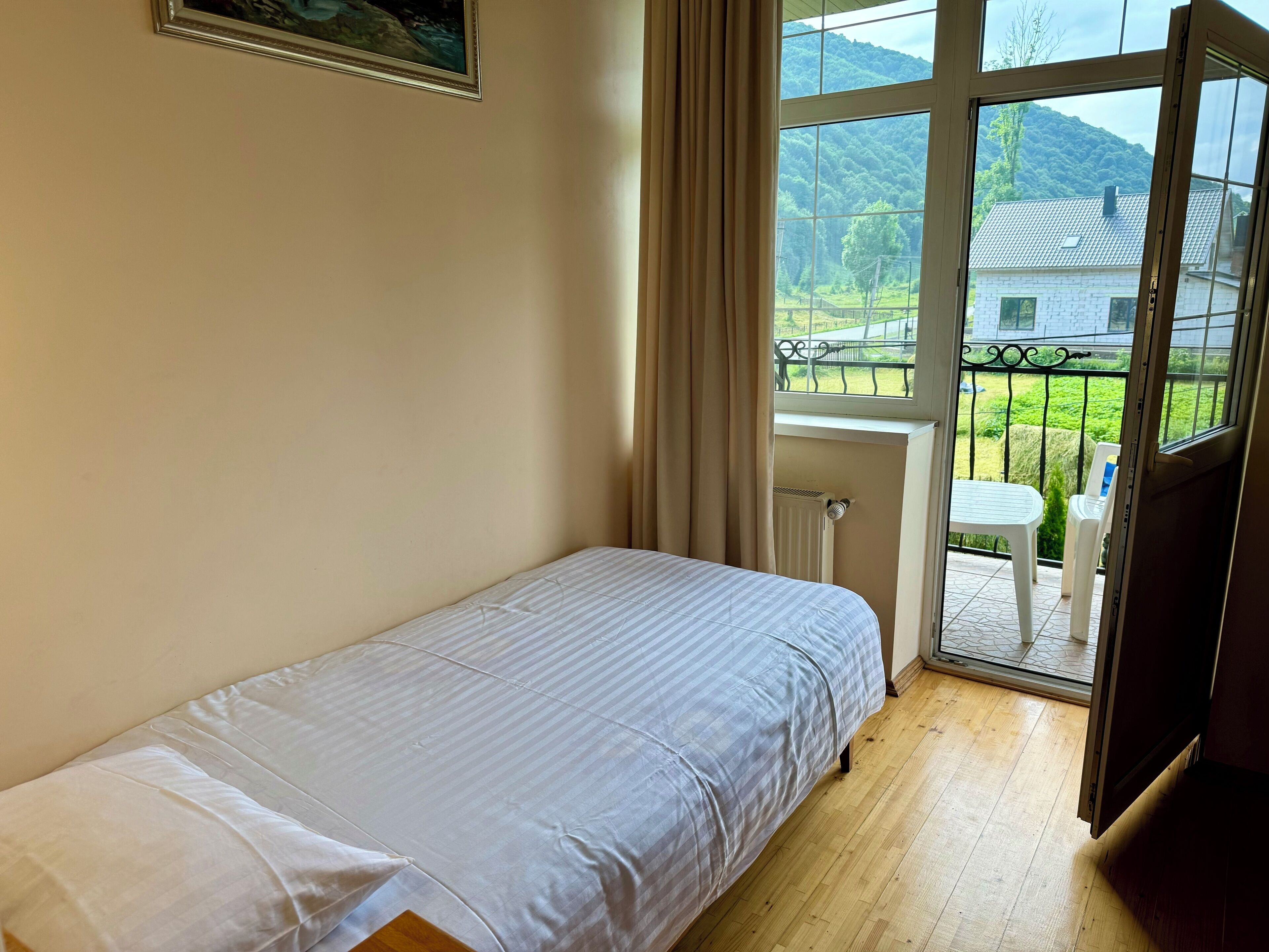 A bedroom with a single bed, a window with curtains, and a view of a green landscape.