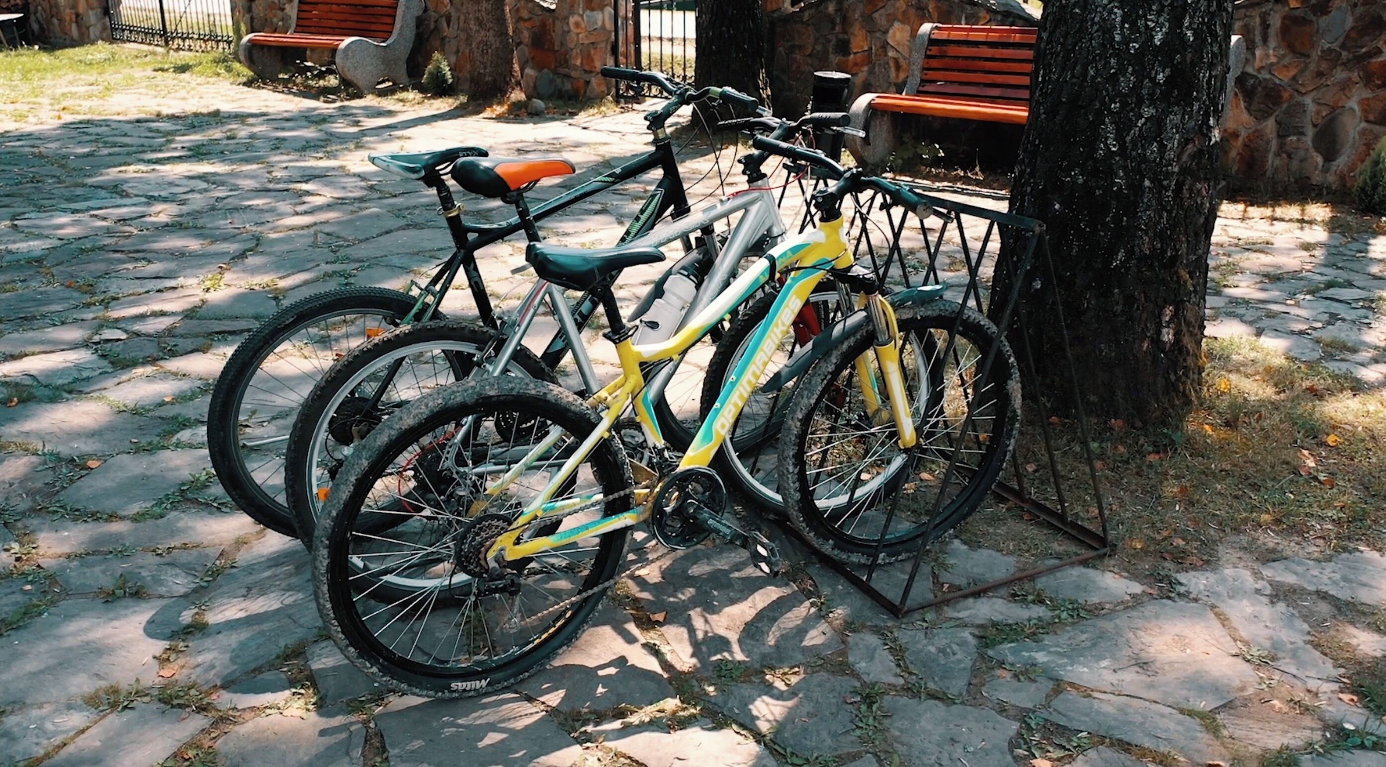 Uma bicicleta estacionada em um caminho de paralelepípedos, com um suporte de metal, cercada por muros de pedra e bancos.
