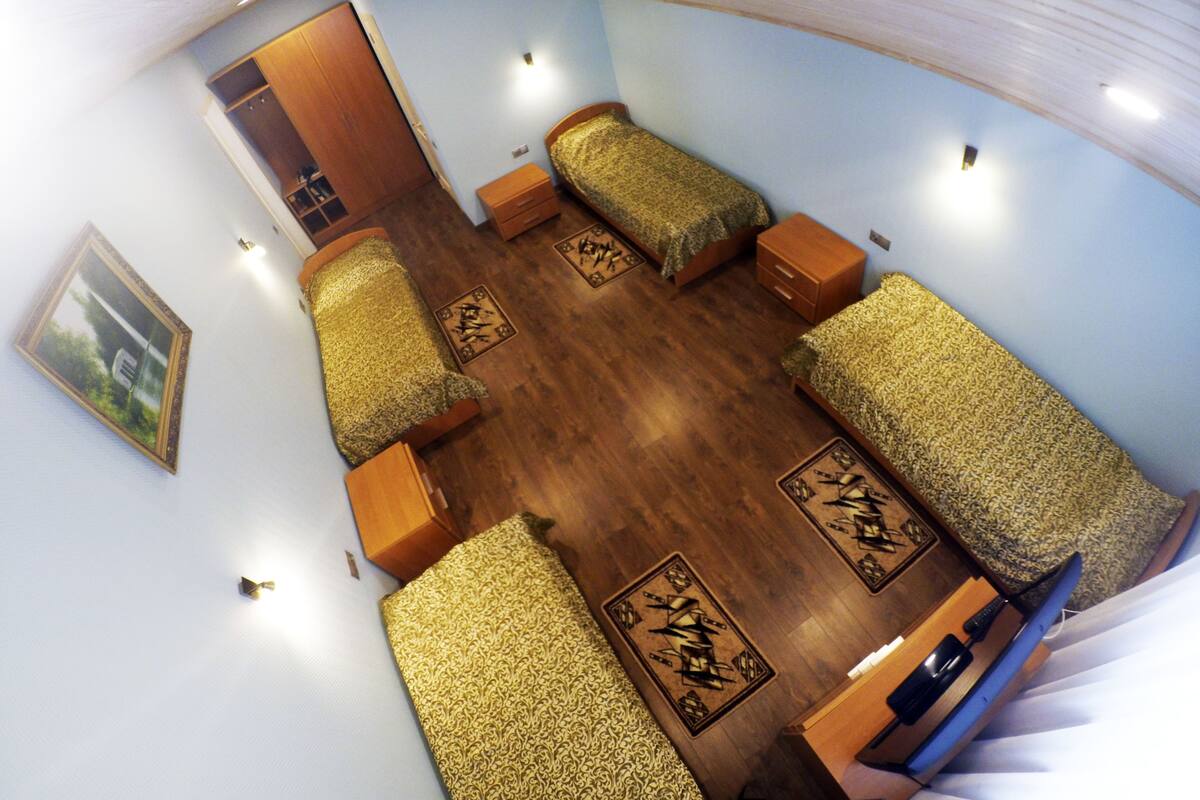 A room with four beds arranged in a U-shape, a wooden wardrobe, a small table, and a wall-mounted picture.