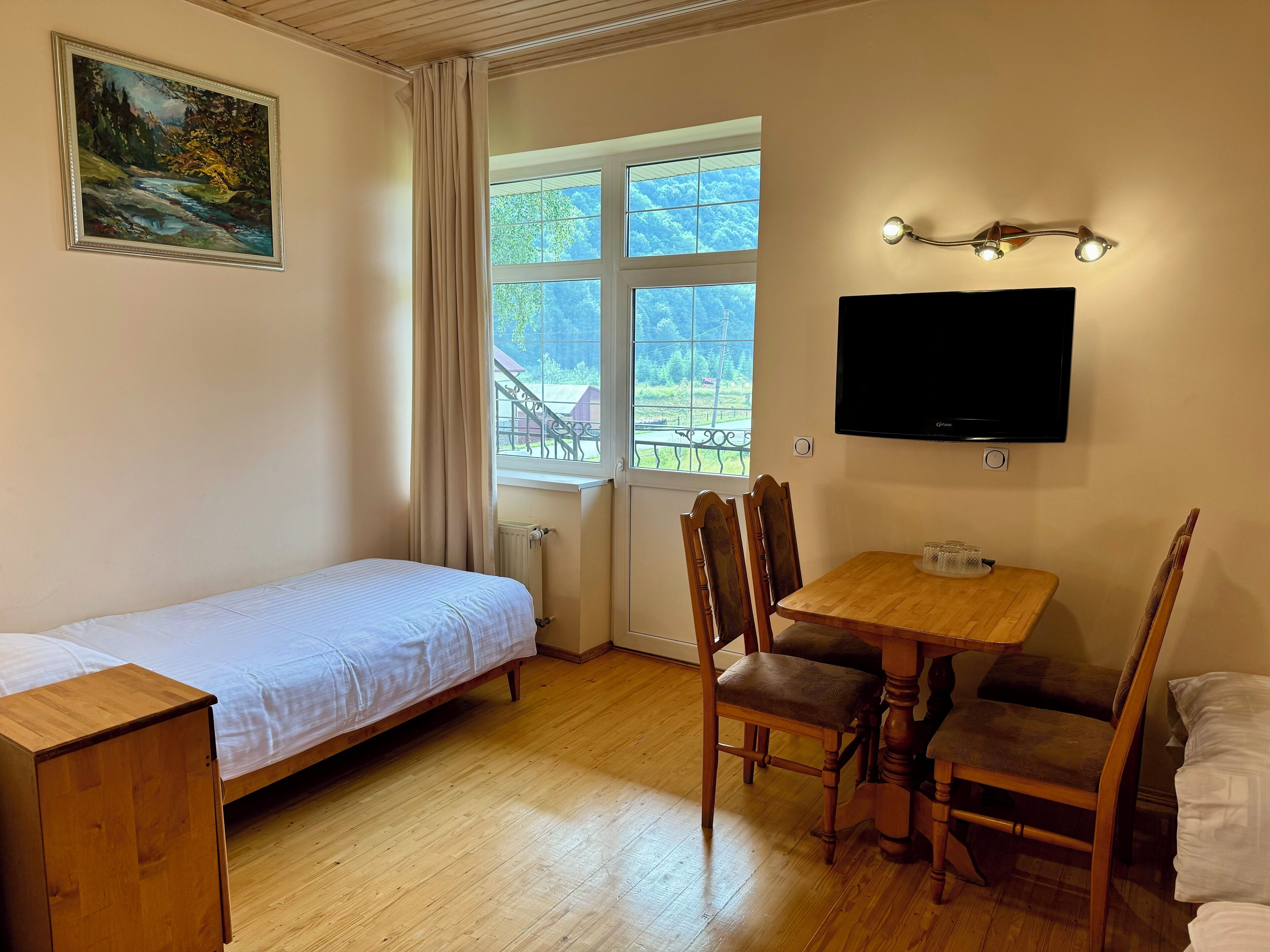 Et værelse med seng, et træbord med stole, et fjernsyn ophængt på væggen og et vindue med gardiner.
