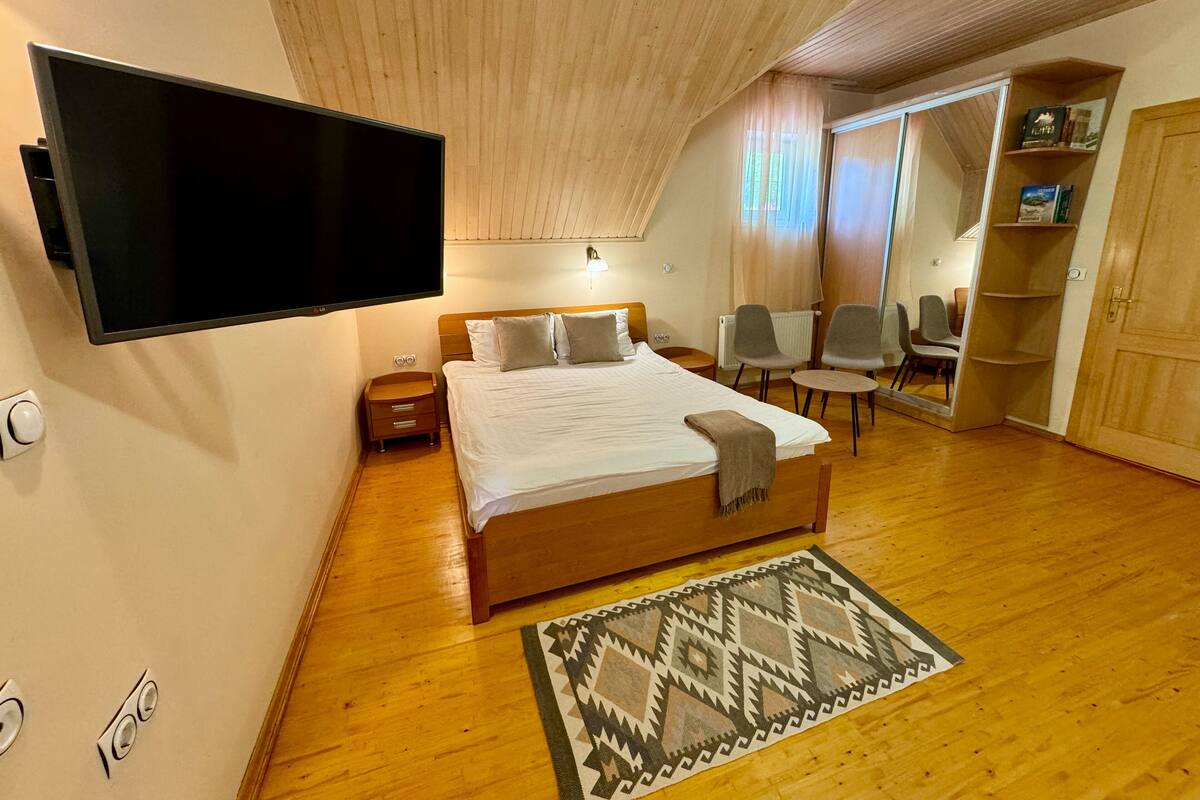 A bedroom with a large bed, a flat-screen TV mounted on the wall, a wooden ceiling, and a view of the adjacent room through a glass door.