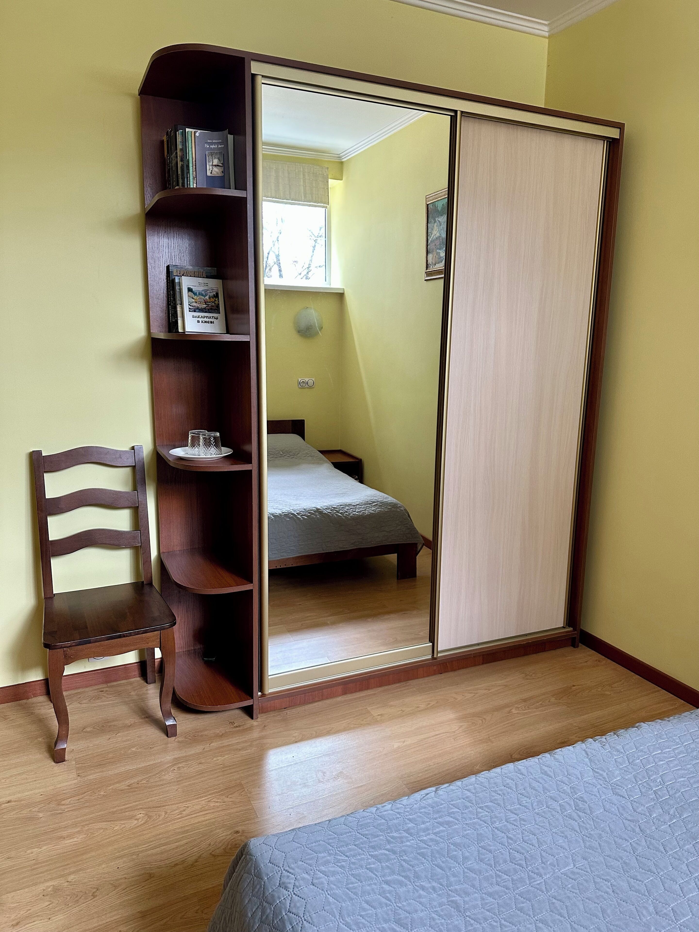 Une chambre à coucher avec un lit, une chaise en bois, une armoire avec un miroir et une étagère remplie de livres.