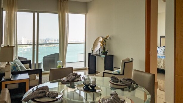 Una habitación de hotel moderna con una mesa de comedor de vidrio, asientos cómodos, vista al mar y una planta pequeña en un estante.