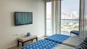 Habitación de hotel con dos camas, televisor, banco y ventana grande con vista a la ciudad.