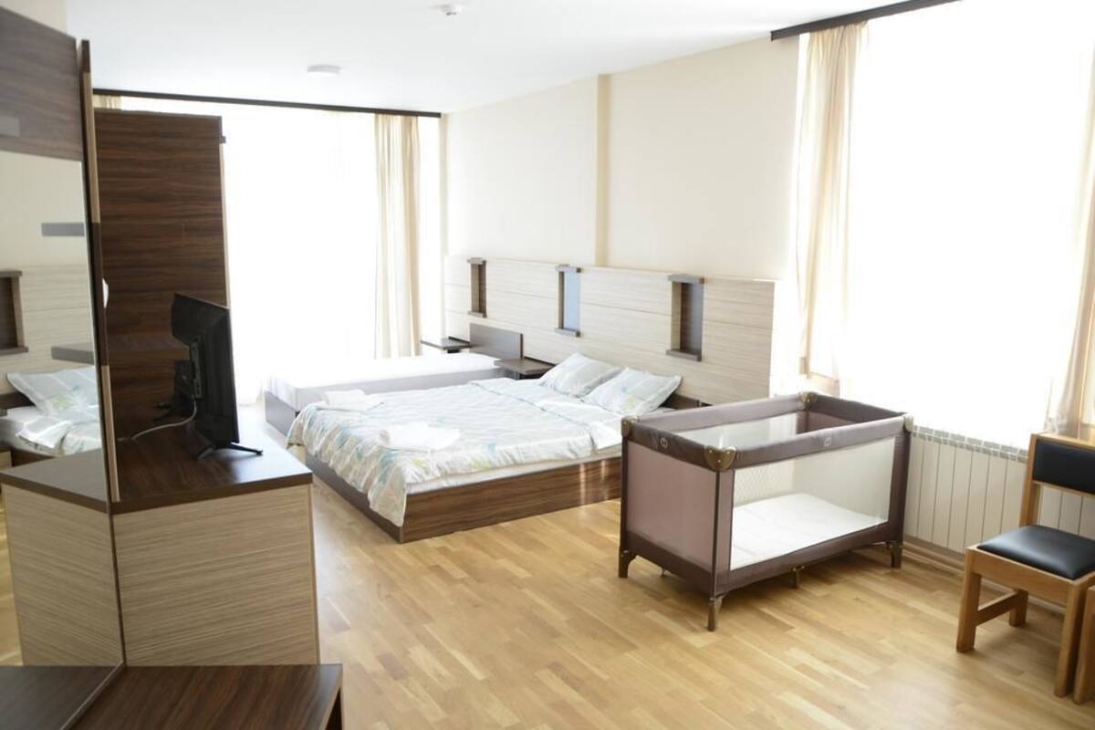 Una habitación de hotel moderna con suelo de madera, una cama con cubrecama blanco, un escritorio con computadora, una silla y una mesita.