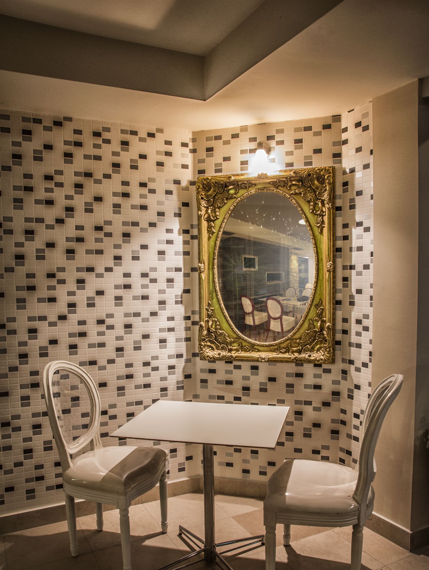 Quarto de canto com uma mesa branca e duas cadeiras brancas, espelho com moldura dourada na parede, e padrão de mosaico em azulejos.