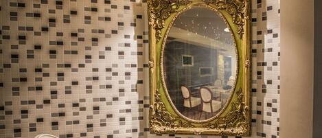 Quarto de canto com uma mesa branca e duas cadeiras brancas, espelho com moldura dourada na parede, e padrão de mosaico em azulejos.