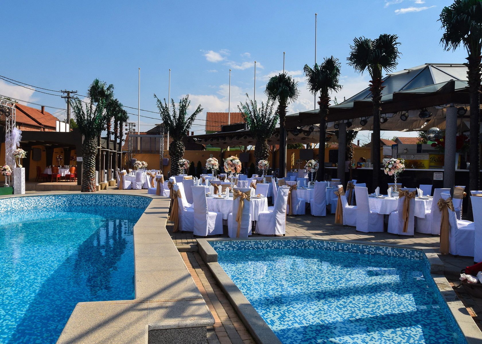 Une cérémonie de mariage organisée au bord de la piscine, avec des tables et des chaises blanches, des palmiers et un ciel d’un bleu éclatant.
