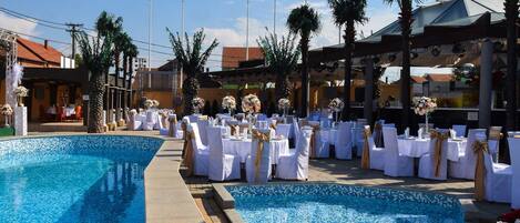 Une cérémonie de mariage organisée au bord de la piscine, avec des tables et des chaises blanches, des palmiers et un ciel d’un bleu éclatant.