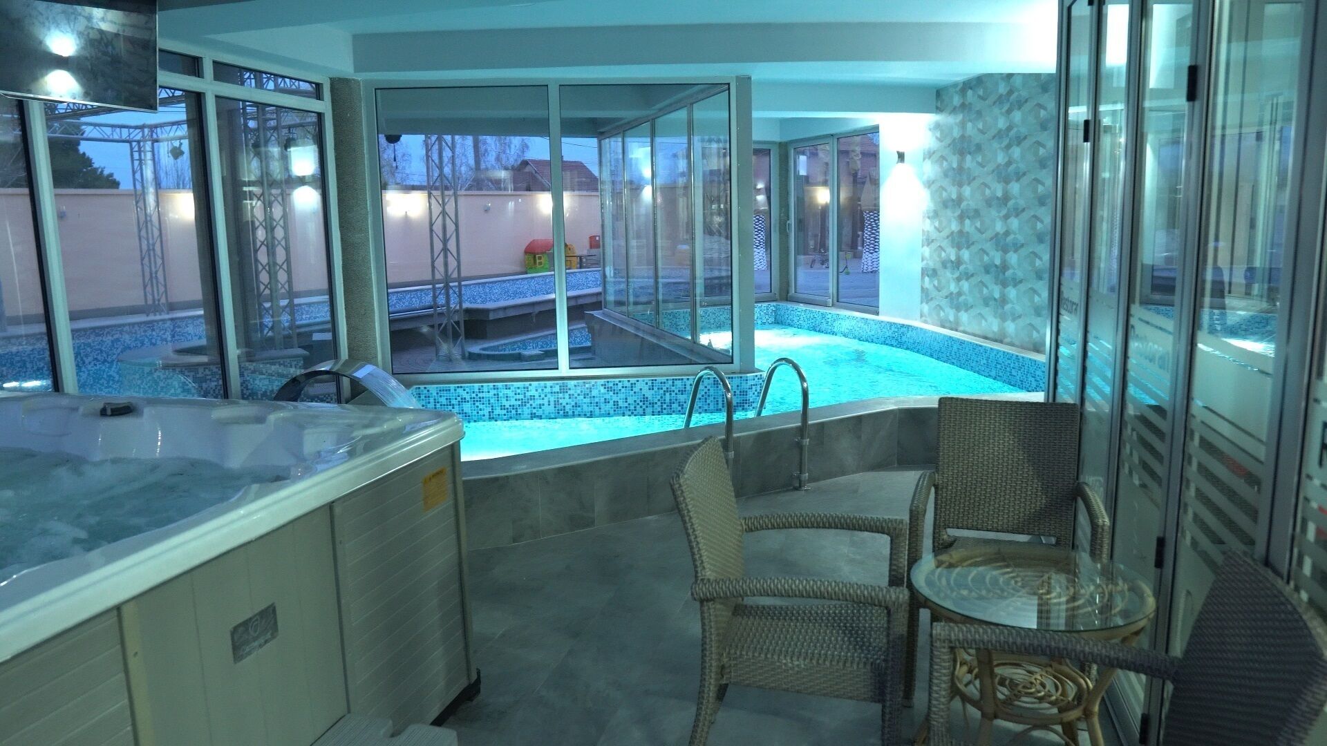 Un área de spa con un jacuzzi, dos sillones de mimbre y una piscina con cerramiento de vidrio.