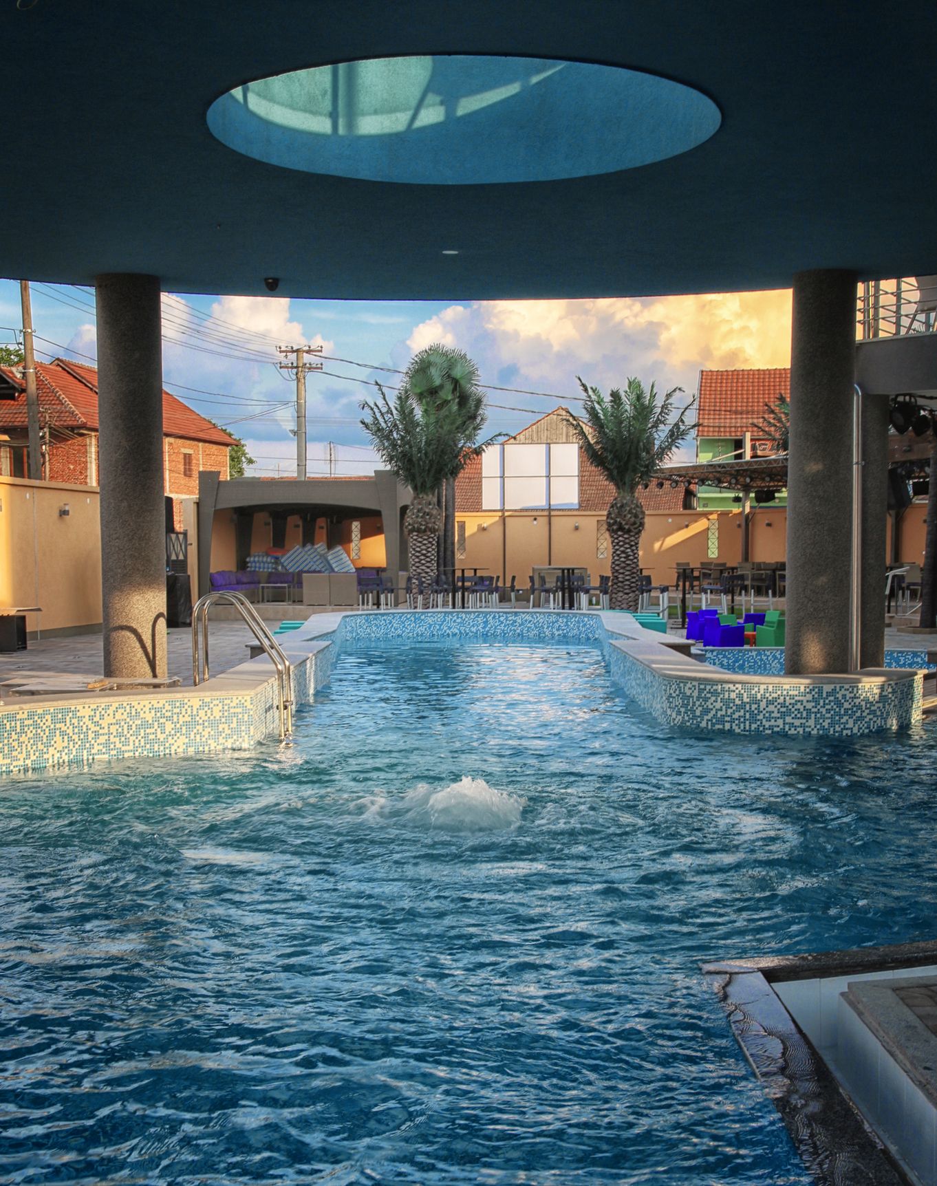 A swimming pool with a tiled edge, palm trees, and a covered seating area.