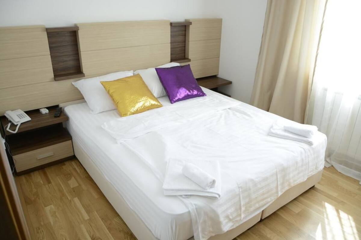 Una cama bien hecha con ropa blanca, dos almohadas doradas y púrpuras, y una toalla blanca al lado.
