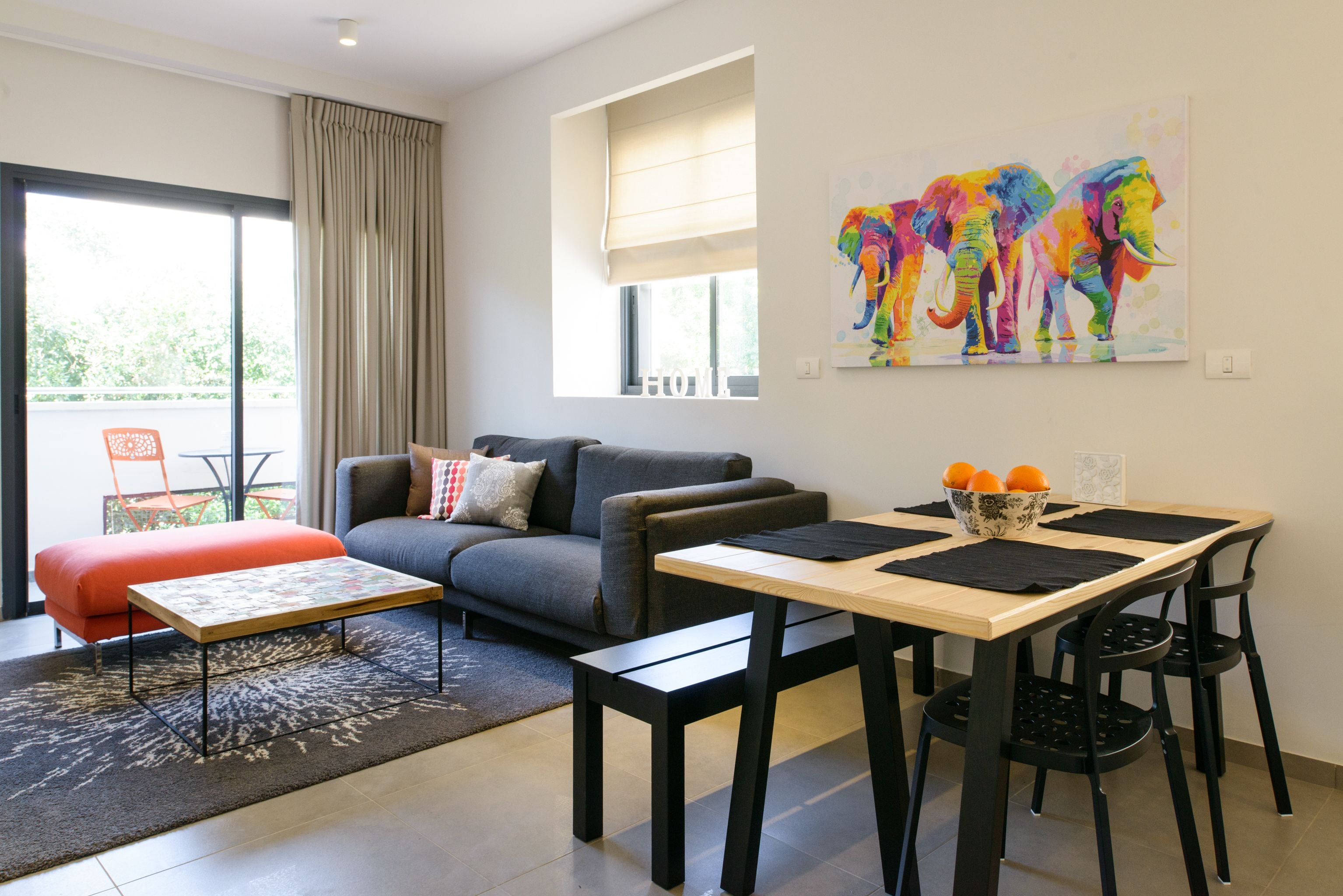 A modern living room with a grey sofa, a wooden coffee table, and a dining area with a table and chairs. There is a colorful elephant painting on the wall and a window with curtains.