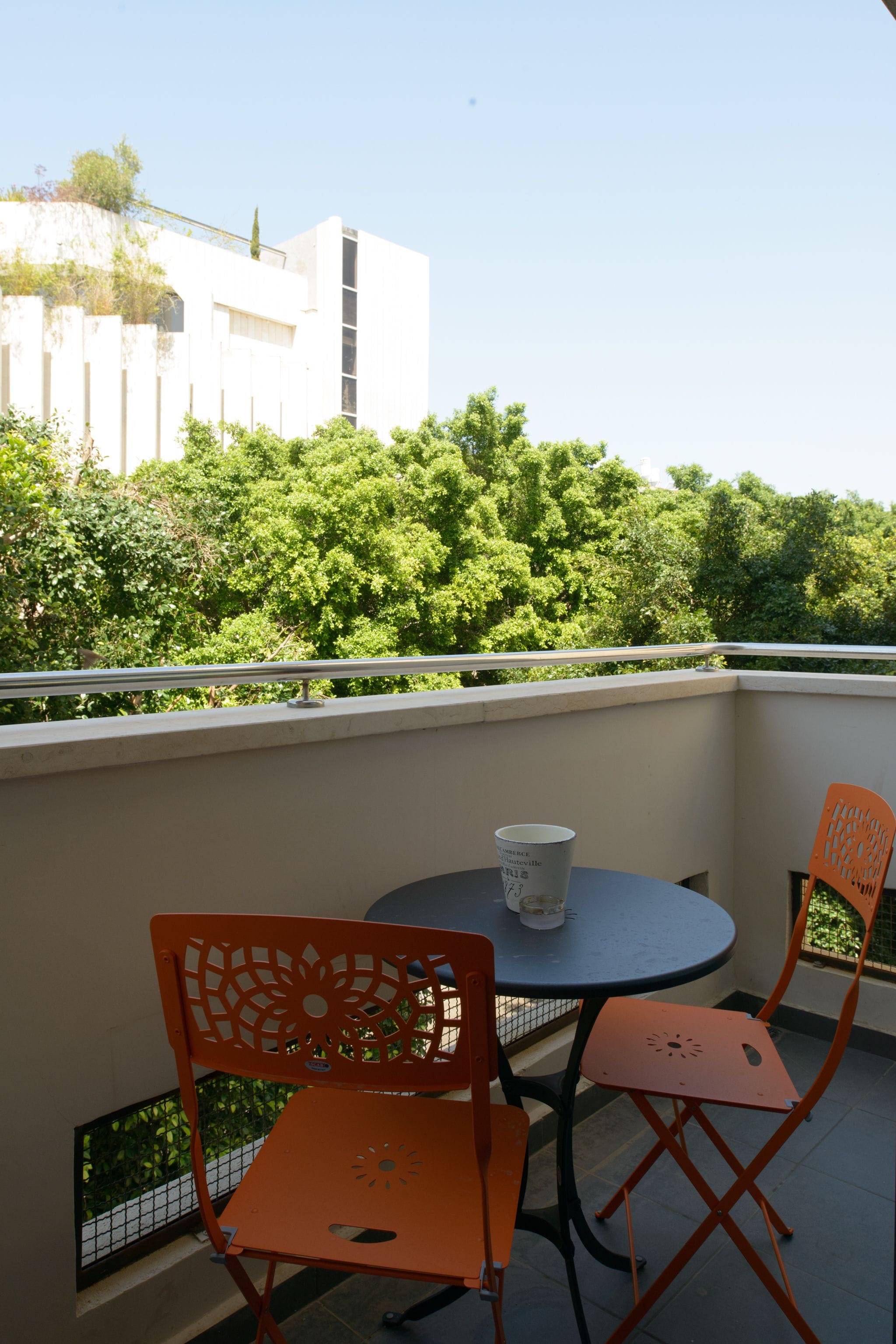 Ein Balkon mit zwei orangefarbenen Stühlen und einem Tisch, mit Blick auf eine üppige Grünfläche und ein weißes Gebäude.