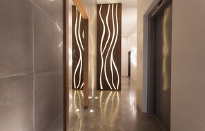 A modern hallway with reflective floor tiles, a wooden panel with light patterns, and a metallic elevator door. - Sunny Luxary Apartments (Tel Aviv)