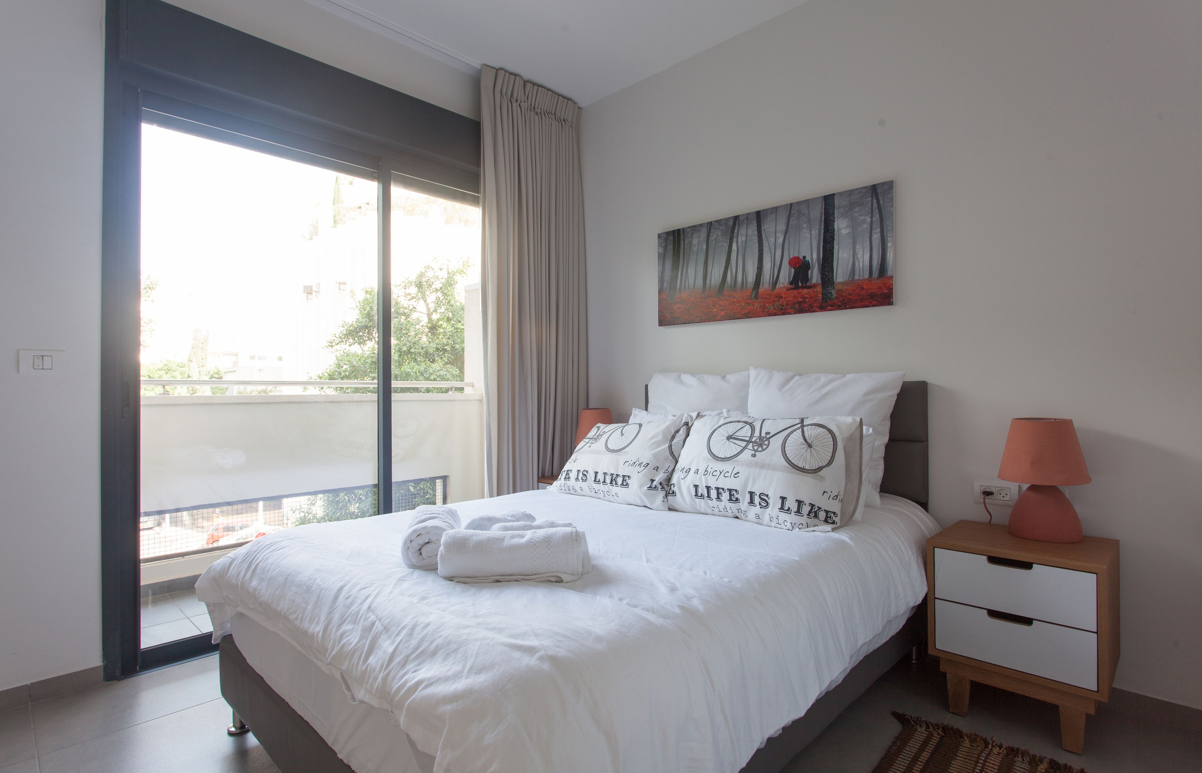 Una camera da letto con un'ampia finestra, un letto con biancheria bianca, un quadro appeso al muro e un comodino con una lampada.
