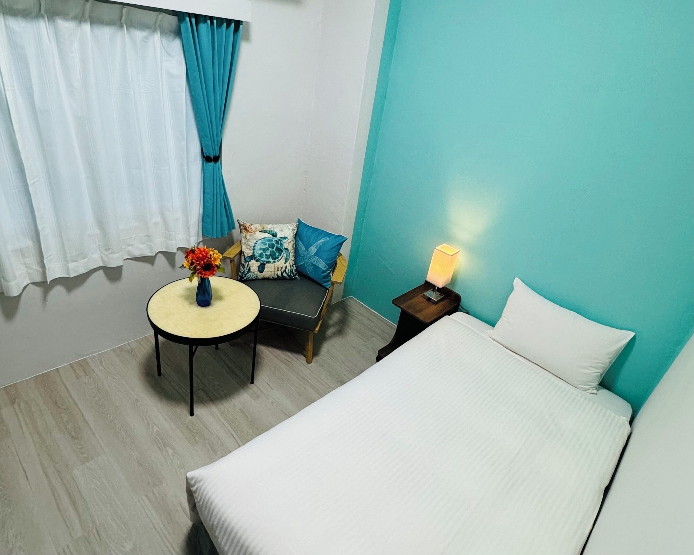 Une chambre d’hôtel avec un lit, une petite table ronde ornée d’un vase de fleurs, une chaise avec un coussin en forme de tortue et une table de chevet avec une lampe.