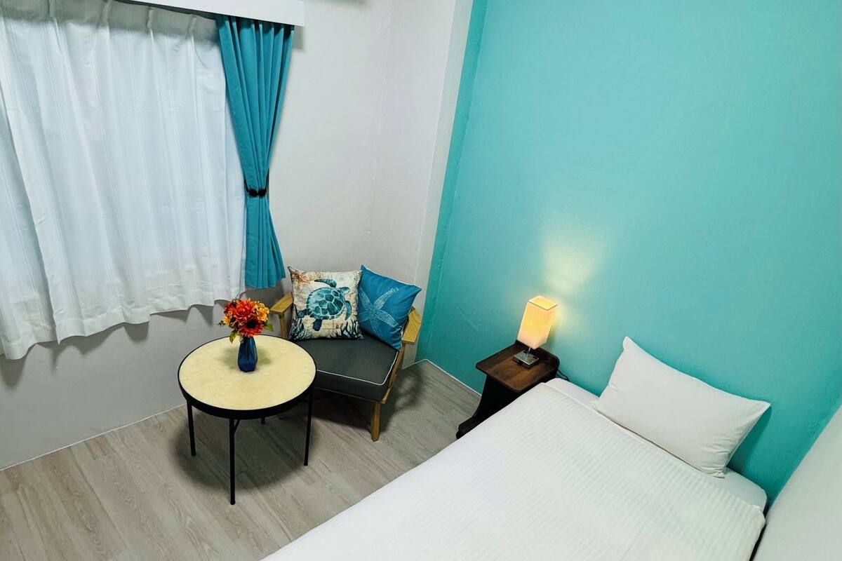 Ein Hotelzimmer mit einem Bett, einem kleinen runden Tisch mit Blumen in einer Vase, einem Sessel mit Kopfkissen in Form einer Schildkröte und einem Nachttisch mit einer Lampe.