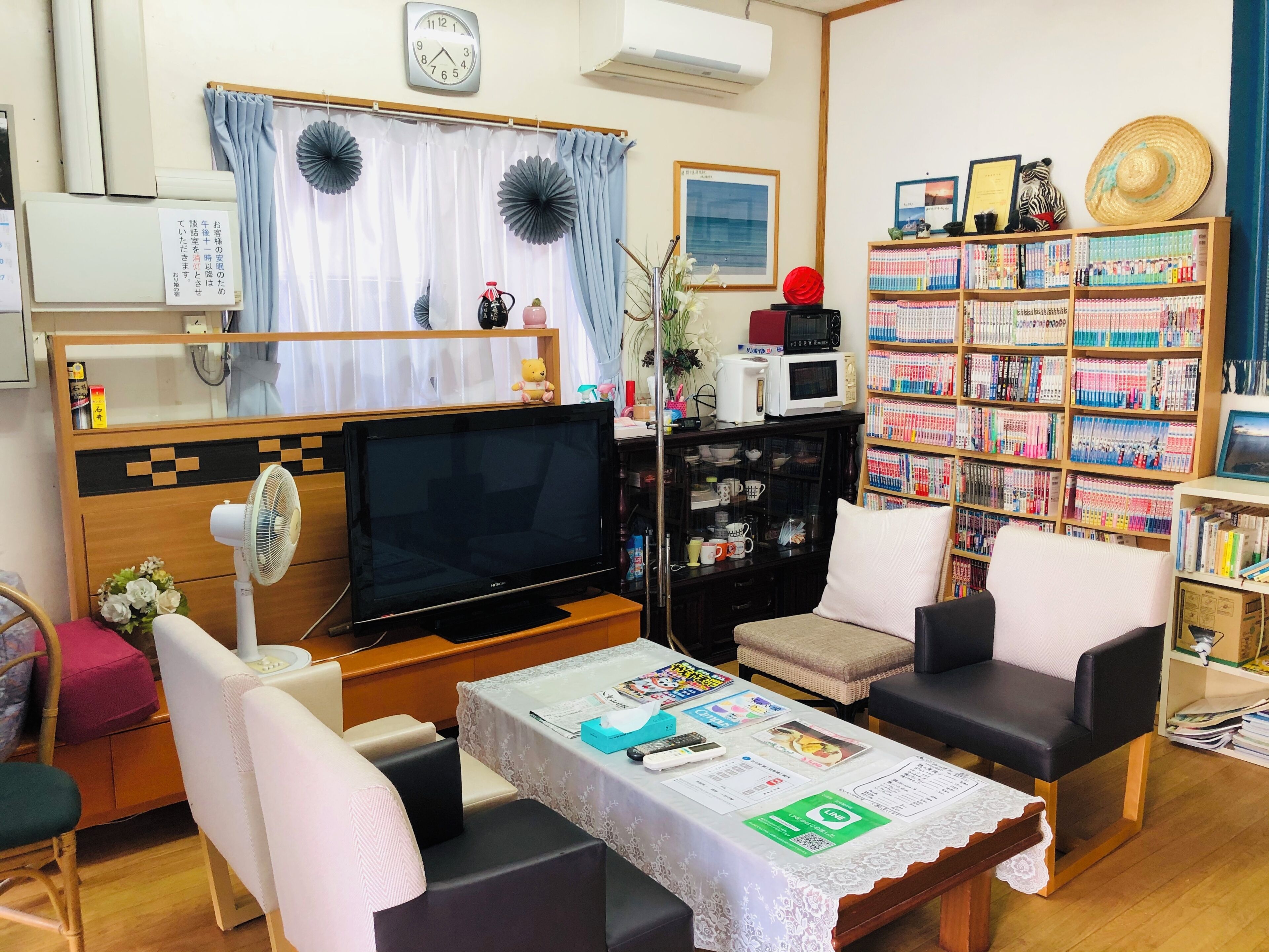 Uma sala de estar com televisão, sofá, cadeiras, uma mesa com papéis, estante e relógio na parede.