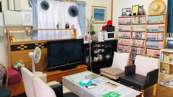 Uma sala de estar com televisão, sofá, cadeiras, uma mesa com papéis, estante e relógio na parede.